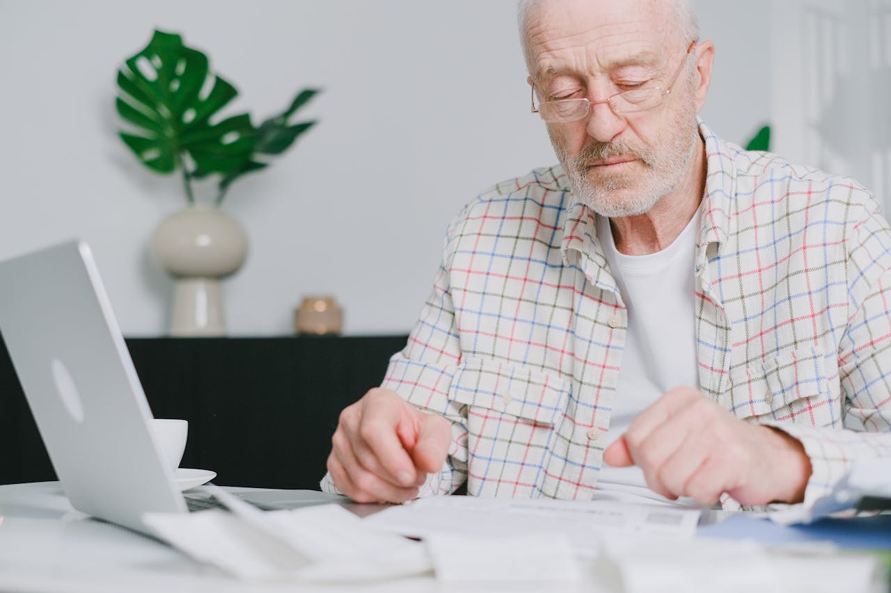 Elderly man with eyeglasses focused on paperwork at home, using laptop.